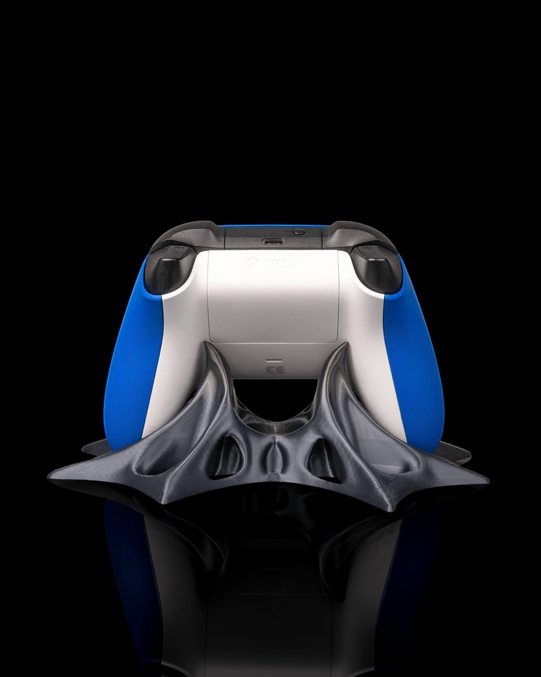 MJR Crafted modern sculptural controller stand in dark grey PLA, holding a white and blue Xbox controller. Black background.